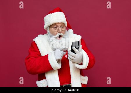 Surprised Santa Claus using smart phone isolated on red background. Stock Photo