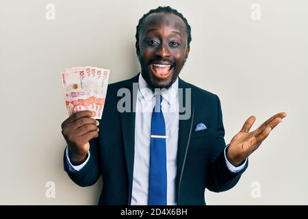 Handsome young black man wearing business suit holding 10 colombian pesos celebrating achievement with happy smile and winner expression with raised h Stock Photo