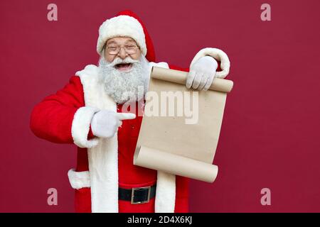 Happy Santa Claus pointing at blank empty wish list isolated on red background. Stock Photo
