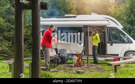 Three Caucasian Friends in Their Late 30s, One Men and Two Woman Having Fun on Wilderness Camping with Modern RV Camper Van. Grilling Bread and Polish Stock Photo