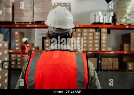 close up of male worker wearing hardhat and orange overalls walking to packages in warehouse  Stock Photo