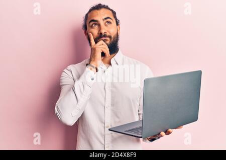 Young arab man holding laptop serious face thinking about question with hand on chin, thoughtful about confusing idea Stock Photo