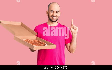 Young handsome man holding delivery pizza cardboard box surprised with an idea or question pointing finger with happy face, number one Stock Photo