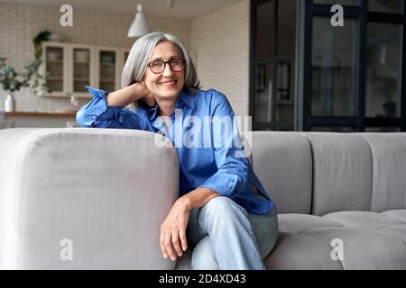 Happy relaxed mature old woman wears glasses sitting on couch at home, portrait. Stock Photo