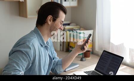Smiling young man working on computer, reading message on phone. Stock Photo