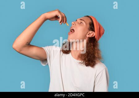 Freestyle. Curly-haired guy wearing hat standing isolated on blue eating fries playful Stock Photo