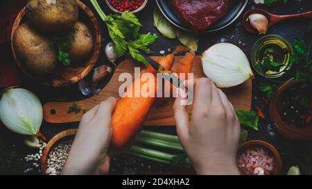 Womens hands are cleaning carrots. Cooking food background. Fresh organic vegetables, ingredients, spices and meat for soup on vintage kitchen table w Stock Photo