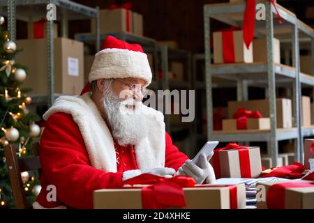 Happy Santa Claus holding cell phone using mobile app sitting at workshop table. Stock Photo