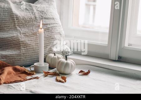Autumn still life. Burning candle in white ceramic candleholder. Dry beech leaves and linen pillow near window. Moody composition with white pumpkins