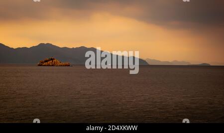 A single little island in the sea surrounded by larger headland an dull clouds in an approaching storm Stock Photo