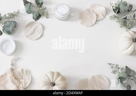 Decorative cosmetics frame. Organic cotton reusable round pads for make up removal. Eucalyptus branches, pumpkins.and moisturizer on white table Stock Photo