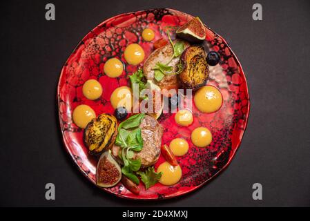 Warm salad, fried pork, fig slices, fried peaches, blueberries, cherry tomato slices, green leaves, pumpkin puree sauce. On a handmade red plate Stock Photo