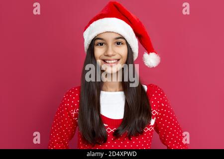 Smiling indian kid girl wear santa hat stand on red background, headshot portrait.