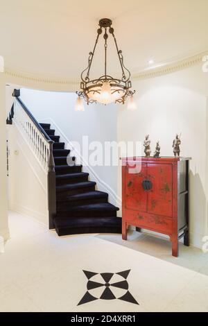 Circular entrance hallway with black arrow marks design inlaid in white marble floor, red antique Chinese cabinet and dark blue carpeted staircase. Stock Photo