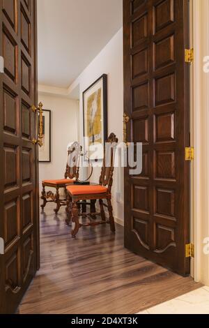 Tall wooden entrance doors, antique orange upholstered wooden high back grid chairs in family room inside contemporary home decorated with paintings. Stock Photo
