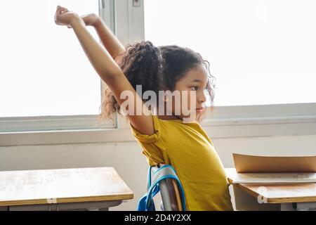 African American school girl stretching with arms raised in classroom, education, learning. Elementary female student taking a break after school. Stock Photo