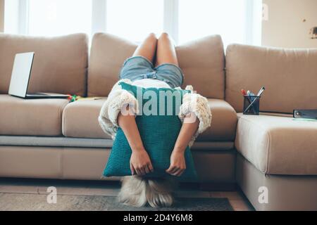 Blonde caucasian woman lying upside-down on a sofa covering face with a pillow feeling tired aftewr working with a laptop Stock Photo