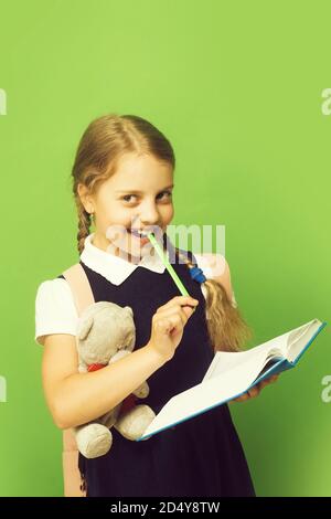 Pupil holds blue book, marker and teddy bear. Kid in school uniform isolated on green background. Study and back to school concept. Girl with braids and happy face. Stock Photo