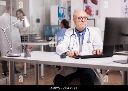 Senior doctor with stethoscope using pc in hospital cabinet while young medic wearing white coat checking report with patient on hospital corridor and nurse in blue uniform. Stock Photo