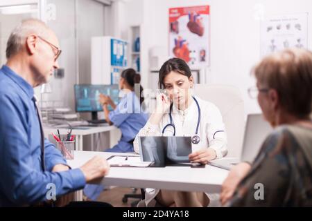 Doctor explaining diagnosis of bone x-ray to pensioner couple and the giving them a bad news. Medic looking at radiography in hospital office Stock Photo