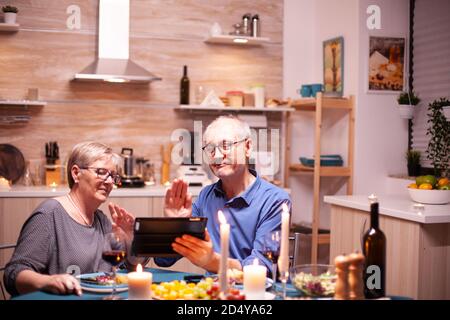 Mature couple saying hello during video conference in kitchen while having romantic dinner using tablet pc. Couple sitting at the table, browsing, talking, using internet,in dining room. Stock Photo