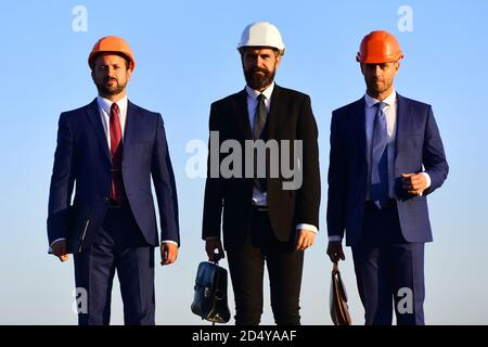 Coworking and construction business concept. Workers and engineer hold meeting outdoors. Foremen present teamwork. Architects with confident faces in suits and helmets hold bags on blue sky background Stock Photo