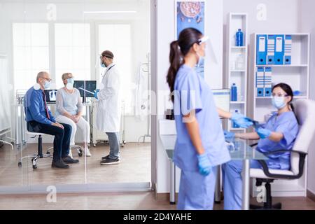 Medical staff wearing face mask against coronavirus in hospital hallway and doctor having a discussion with old couple in examination room. Assistant working on reception computer. Stock Photo
