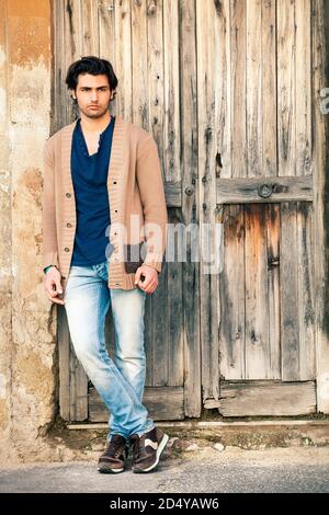 Handsome young man is leaning against the wall near the door of an old house entrance. A ruined wooden door. Stock Photo