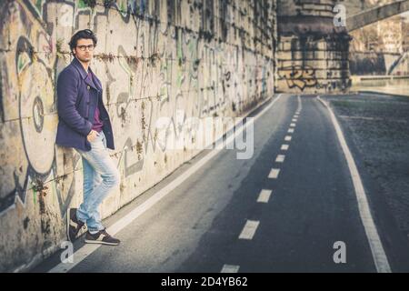 Handsome and charming young man outdoors leans against a wall in the city. Dressed in fashionable clothes and wearing eyeglasses. Coat and jeans. Stock Photo