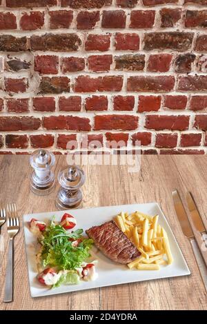 Rustic wooden table with a set of silverware , White plate with Beef steack, french fries and tomato on salad. Stock Photo