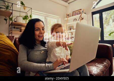 Little toddler girl sitting on mother lap using laptop on couch and pointing on screen showing something Stock Photo