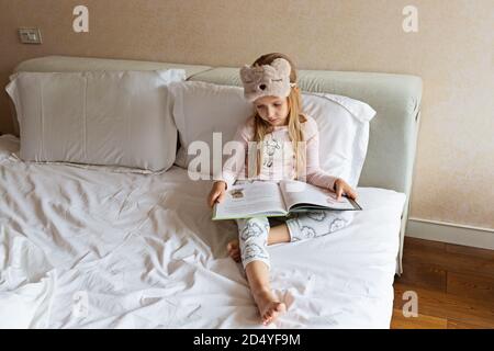 Cute little caucasian girl in pyjama reading a book. Kid sitting on a bed in the room. Stay at home during coronavirus covid-19 lockdown. Stock Photo