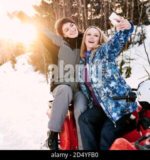 Two cheerful pretty girls posing taking selfie against setting sun in snowy mountains sitting on red quad-bike. Concept of winter entertainment