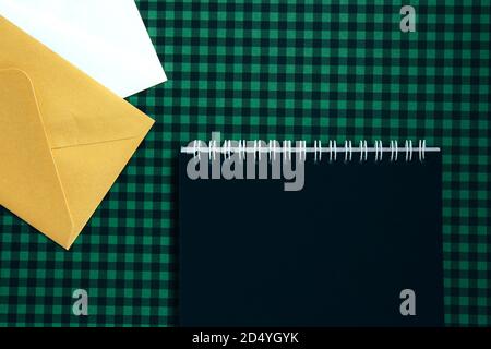 Open spiral notepad with black pages and white and beige envelopes on a green checkered background. List of goals for the new year. Blank template Stock Photo