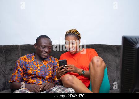 young african couple sitting on a couch smiling while viewing content on a phone together Stock Photo