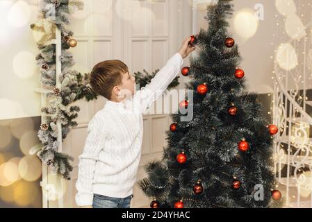 Cute little boy decorates the Christmas tree with New Year's toys and red balls. A boy in a white knitted sweater sits next to gifts and hangs balls on an artificial spruce. Family Christmas concept. Stock Photo