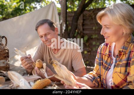 Smiling middle-aged couple sitting at table, shucking corn Stock Photo