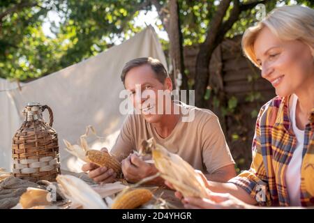 Happy middle-aged couple sitting at table, shucking corn Stock Photo