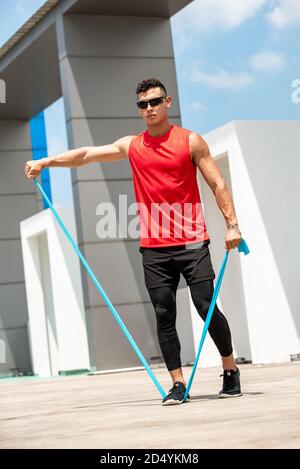 Handsome muscular sportsman doing shoulder lateral raise exercise with resistance band outdoors on rooftop Stock Photo