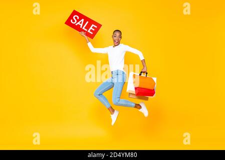 Excited happy African American woman jumping with red sale sign and shopping bags in hands isolated on yellow background Stock Photo