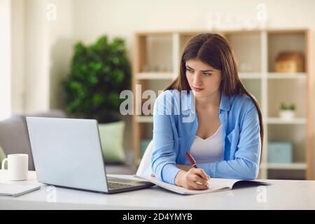 Young female student writing in notebook while watching online lecture sitting at desk at home Stock Photo