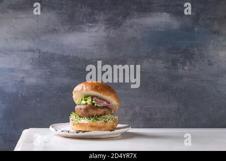 Homemade vegan vegetarian burger in wheat bun with portobello mushroom, avocado salsa and sprouts on white ceramic plate over white marble table. Stock Photo