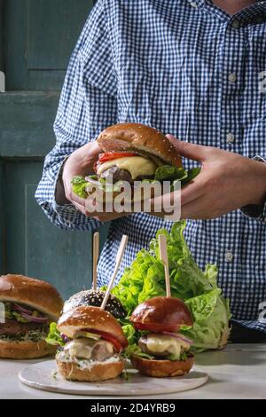 Man in blue shirt holding in hands homemade classic burger in wheat bun with beef cutlets, melted cheese, vegetables. Table with ingredients and mini Stock Photo