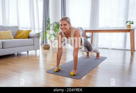 Beautiful active young woman exercise plank pose with dumbbells at home. Side view. Healthy lifestyle and fitness concept. Series of exercise poses. Stock Photo