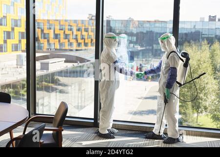 Full length portrait of two disinfection workers wearing hazmat suits shaking hands while standing against window in office, copy space Stock Photo
