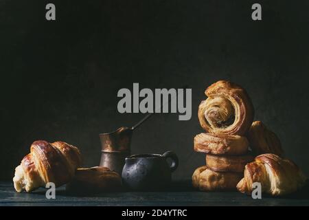 Variety of homemade puff pastry buns cinnamon rolls and croissant served with vintage coffee pot on wooden table. Dark still life. Copy space Stock Photo