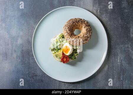Vegetarian whole grain bagel sandwich with chopped avocado, cream cheese, sun dried tomatoes, egg, served on ceramic plate over blue texture backgroun Stock Photo