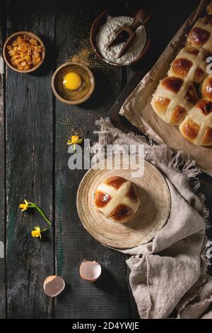 Homemade Easter traditional hot cross buns on plate and oven tray with baking paper and ingredients above over dark wooden background. Top view, space Stock Photo