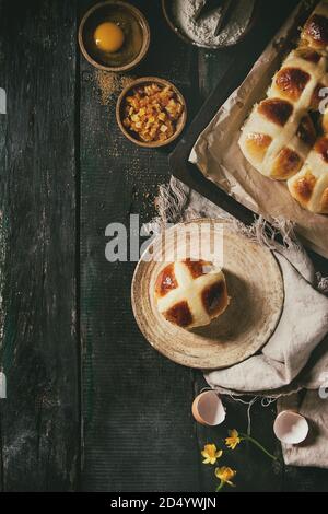 Homemade Easter traditional hot cross buns on plate and oven tray with baking paper and ingredients above over dark wooden background. Top view, space Stock Photo