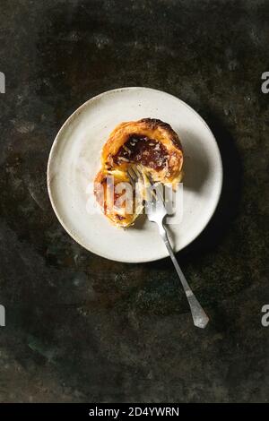 Broken Traditional Portuguese egg tart dessert Pasteis Pastel de nata on ceramic plate with fork over dark metal background. Flat lay, space Stock Photo